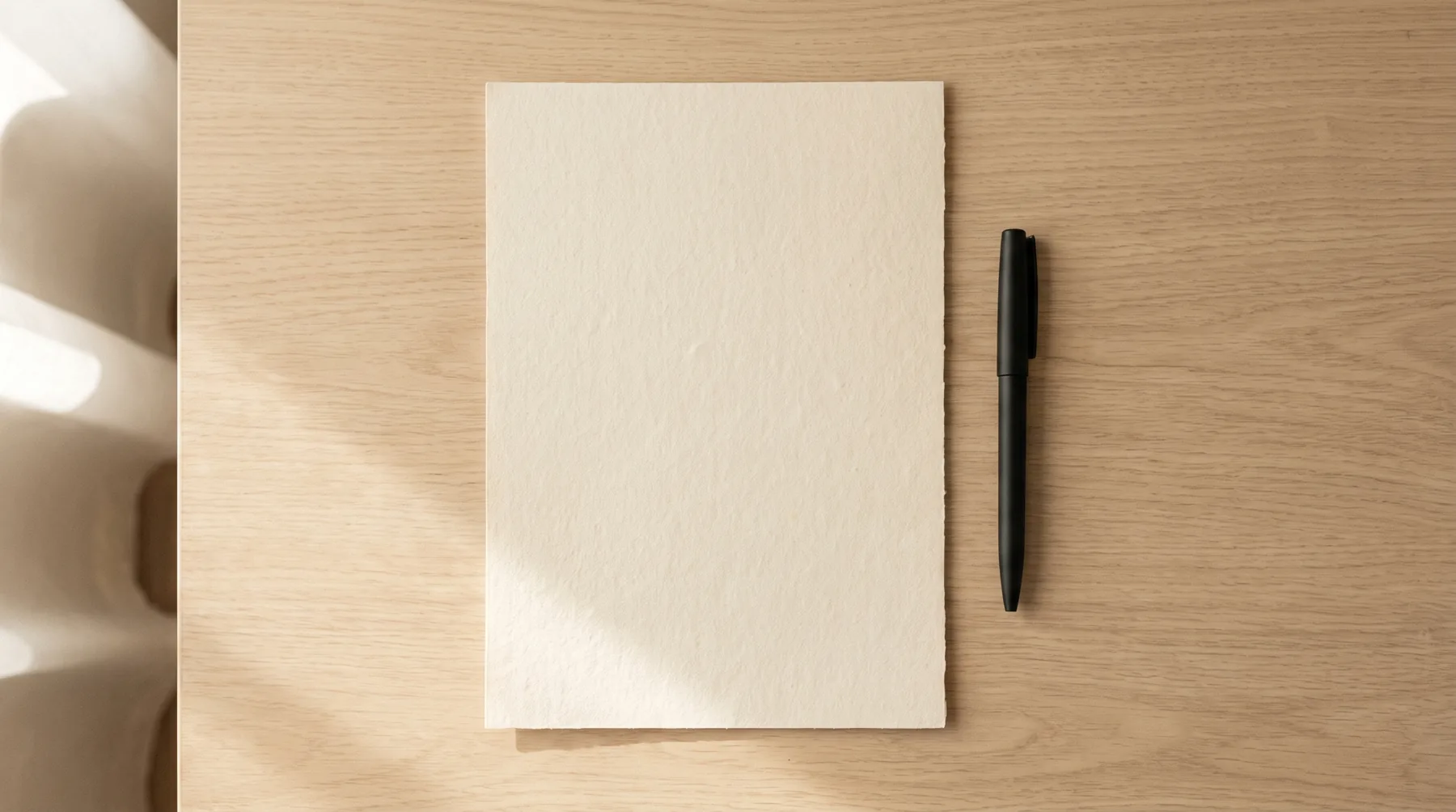 A sheet of cream laid paper and a pen on a pale oak desk in morning light