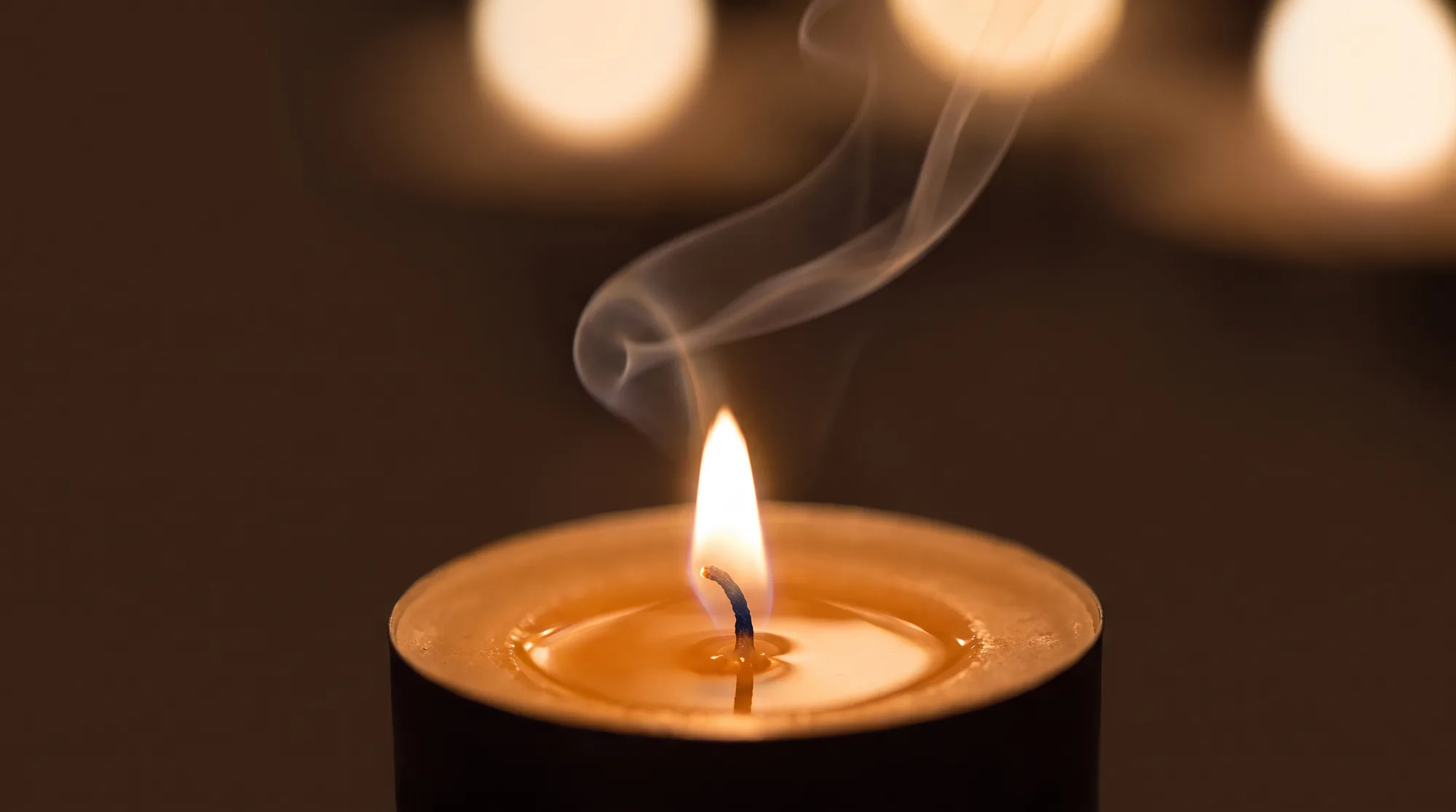 A single tea-light burning against a deep warm brown, a wisp of smoke rising from the wick