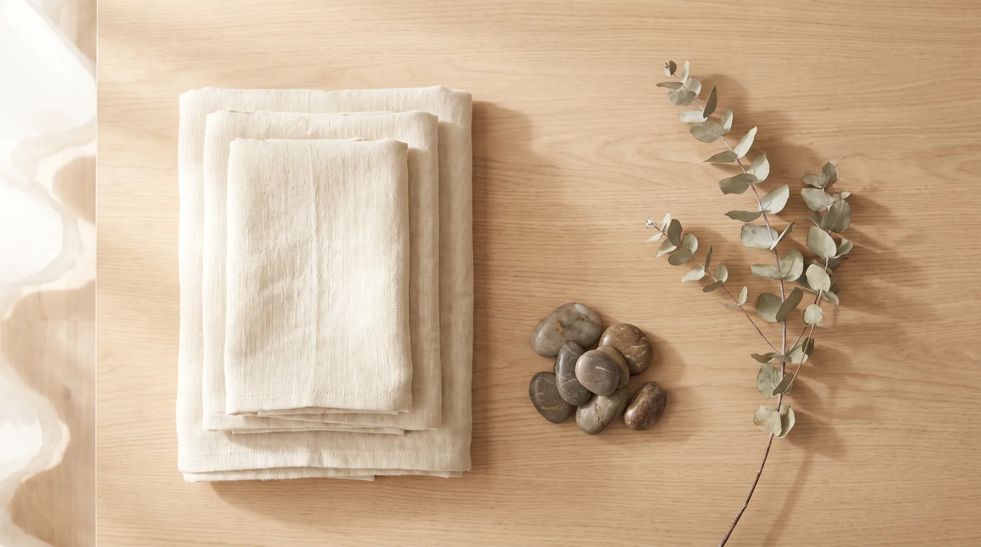 Folded cream linen towels, river stones and a sprig of eucalyptus on pale oak